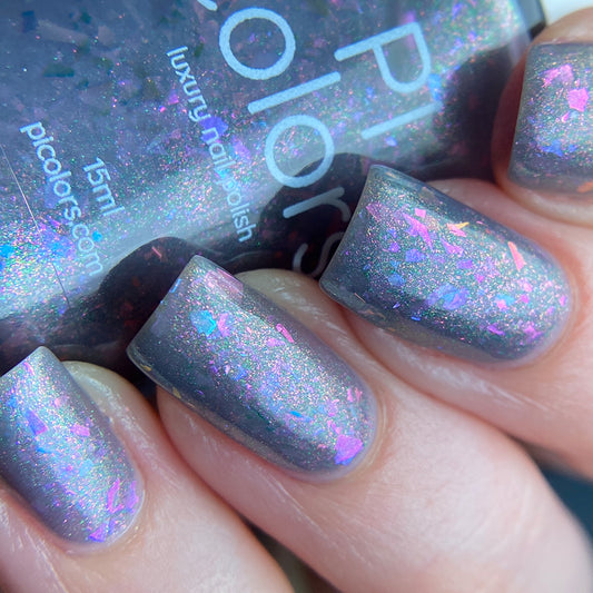 Close-up of a hand with nails painted in "The First Rule.000 Gray Nail Polish" by PI Colors, featuring a luxurious shimmer of gray highlighted with pink and purple iridescent flakies. The nail polish bottle is partially visible in the background.