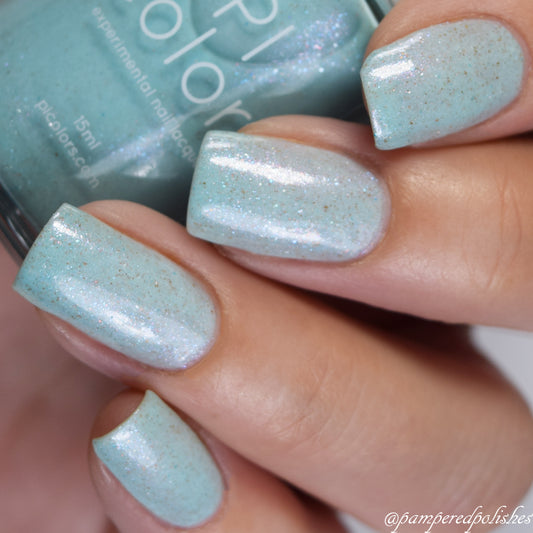 Close-up of a hand adorned with nails coated in Marshmellow Dream.001, a shimmering pale green color-changing nail polish featuring speckles. The glittery finish sparkles under light, while a partially visible bottle of PI Colors Luxury Nail Lacquers is held in the background.