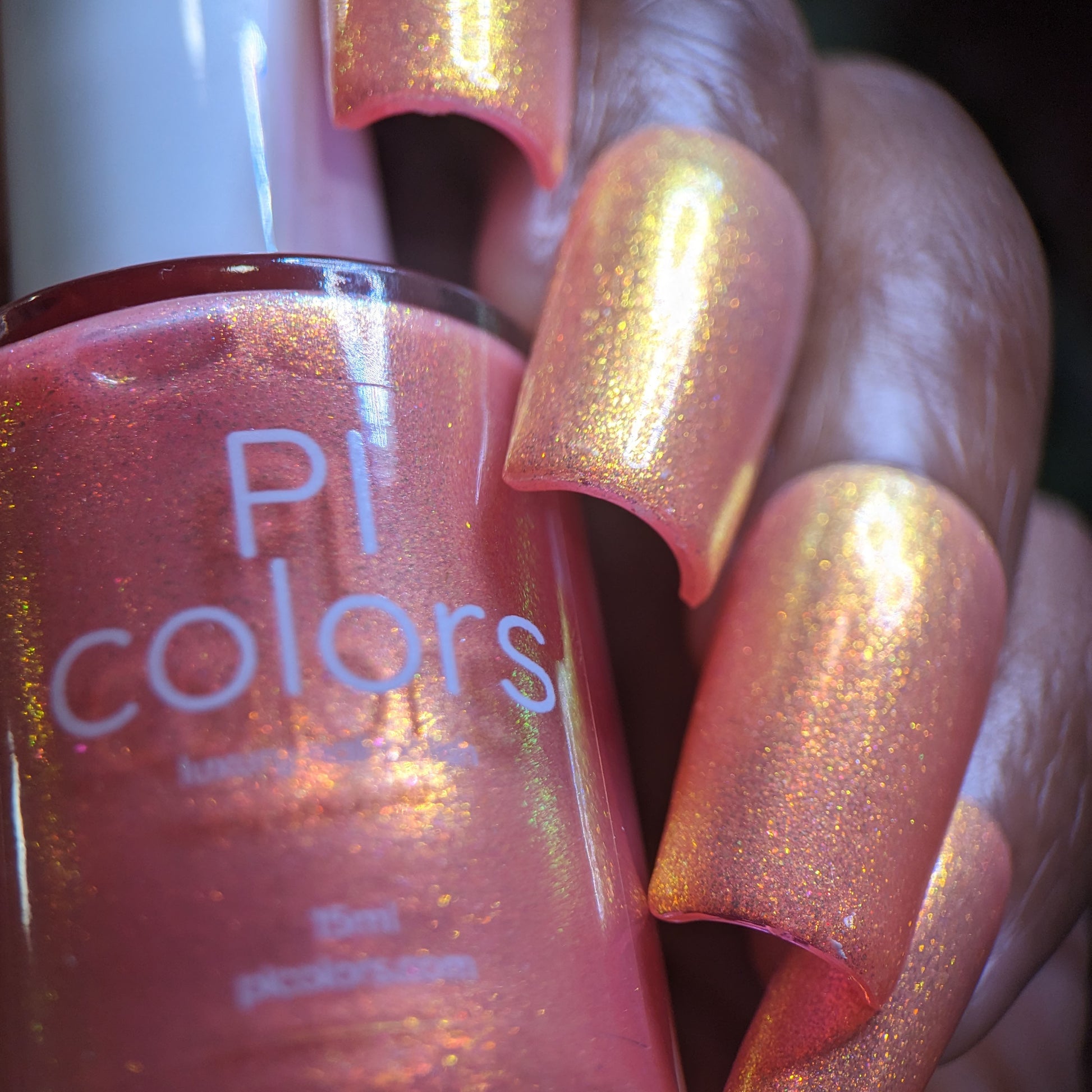 A close-up captures a hand with long, shimmery peach-colored nails holding a bottle of Alstroemeria.000 by PI Colors. The iridescent polish seamlessly complements the nails, creating an elegant look reminiscent of the delicate beauty of Alstroemeria.