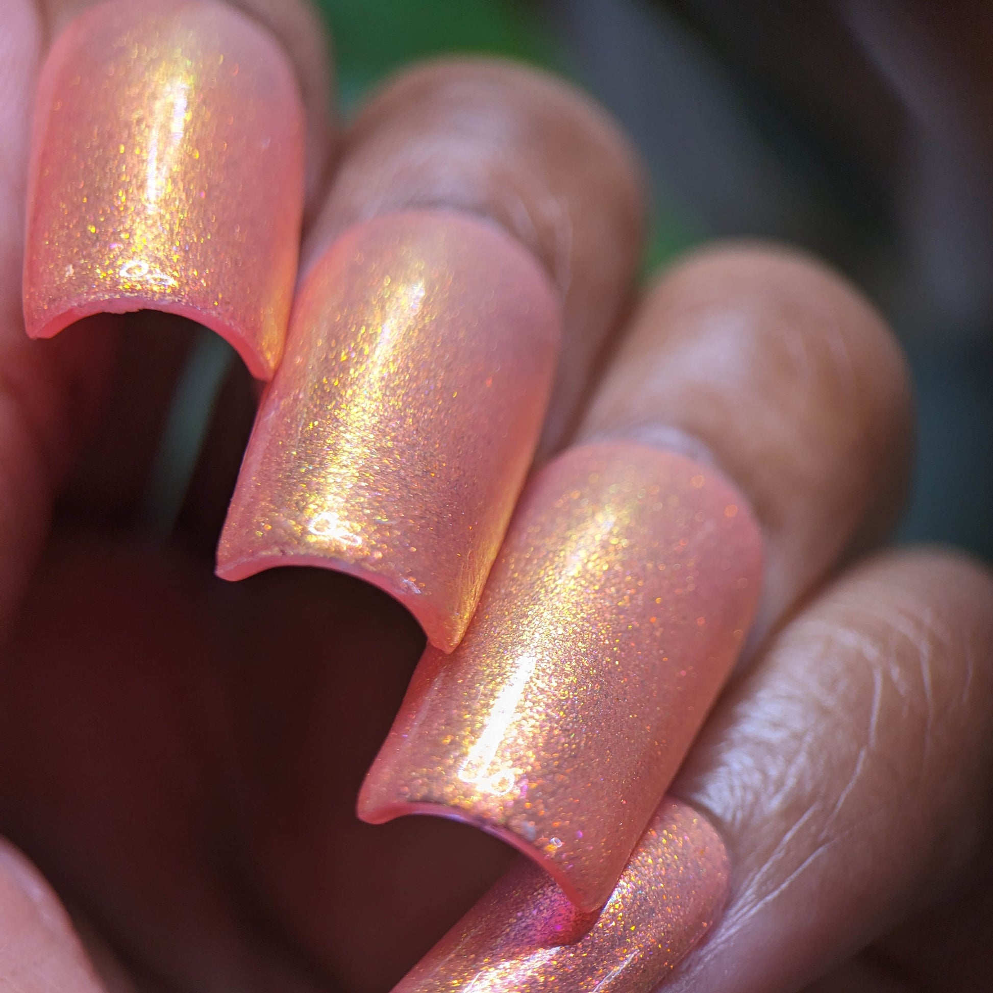 Close-up of a hand showcasing long, pink-orange acrylic nails with a glittery shimmering finish. The iridescent look under bright lighting captures the elegance of PI Colors' Alstroemeria.000 Luxury Nail Lacquer.