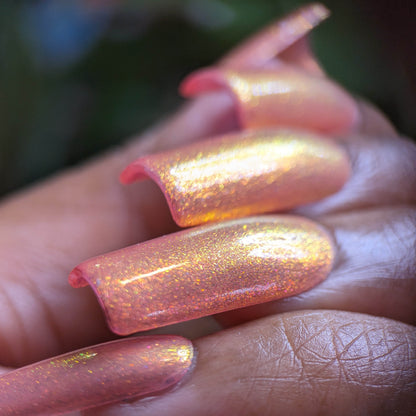 A close-up shot showcases a person's hand with long, shimmering nails featuring a peach and gold holographic design by Luxury Nail Lacquers. The nails capture the light beautifully, displaying a gradient of colors that evoke the essence of PI Colors' Alstroemeria.000 with a glittery finish against a softly blurred background.