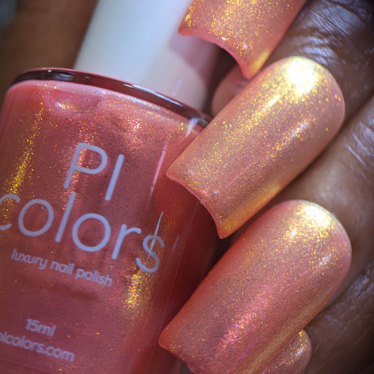 A close-up of a hand with peach-colored nail polish that has a golden shimmer. The Alstroemeria.000 luxury lacquer by PI Colors is held next to the nails, highlighting the perfectly matching shade with its shimmering finish.