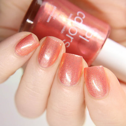 A hand displays nails adorned with shimmery coral polish from PI Colors, featuring Alstroemeria.000. The nails gleam with a glossy finish while the person holds a matching bottle of the polish in the background.