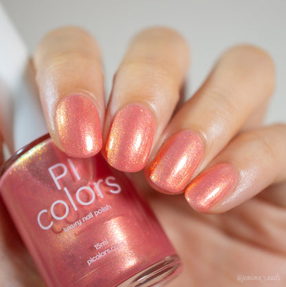 A close-up of a hand with short, rounded nails painted in a shimmering shade of Alstroemeria.000 by PI Colors, with a bottle of this luxury nail lacquer nestled gracefully between the fingers, exuding elegance and style.