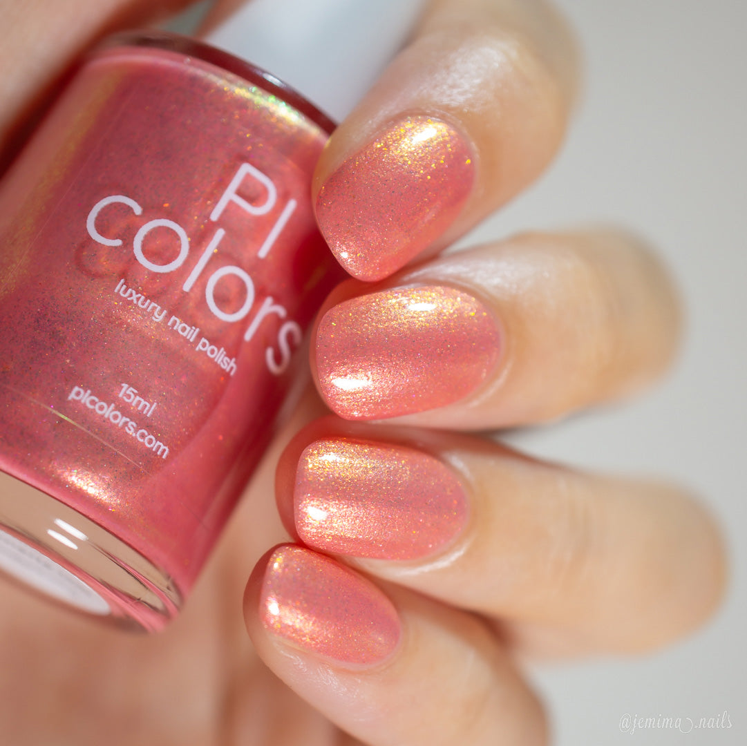 A close-up shot captures a hand with nails adorned in a shimmery coral pink polish from PI Colors, showcasing their Alstroemeria.000 Luxury Nail Lacquer. The bottle displays the same radiant shade, complete with a luxurious finish and subtle gold flecks.
