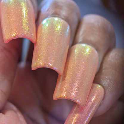 Close-up of a hand with long, square-shaped nails in shimmery peach, enhanced by PI Colors' Luxury Nail Lacquer. The Alstroemeria.000 shade boasts an iridescent quality with hints of pink and gold, capturing light elegantly.