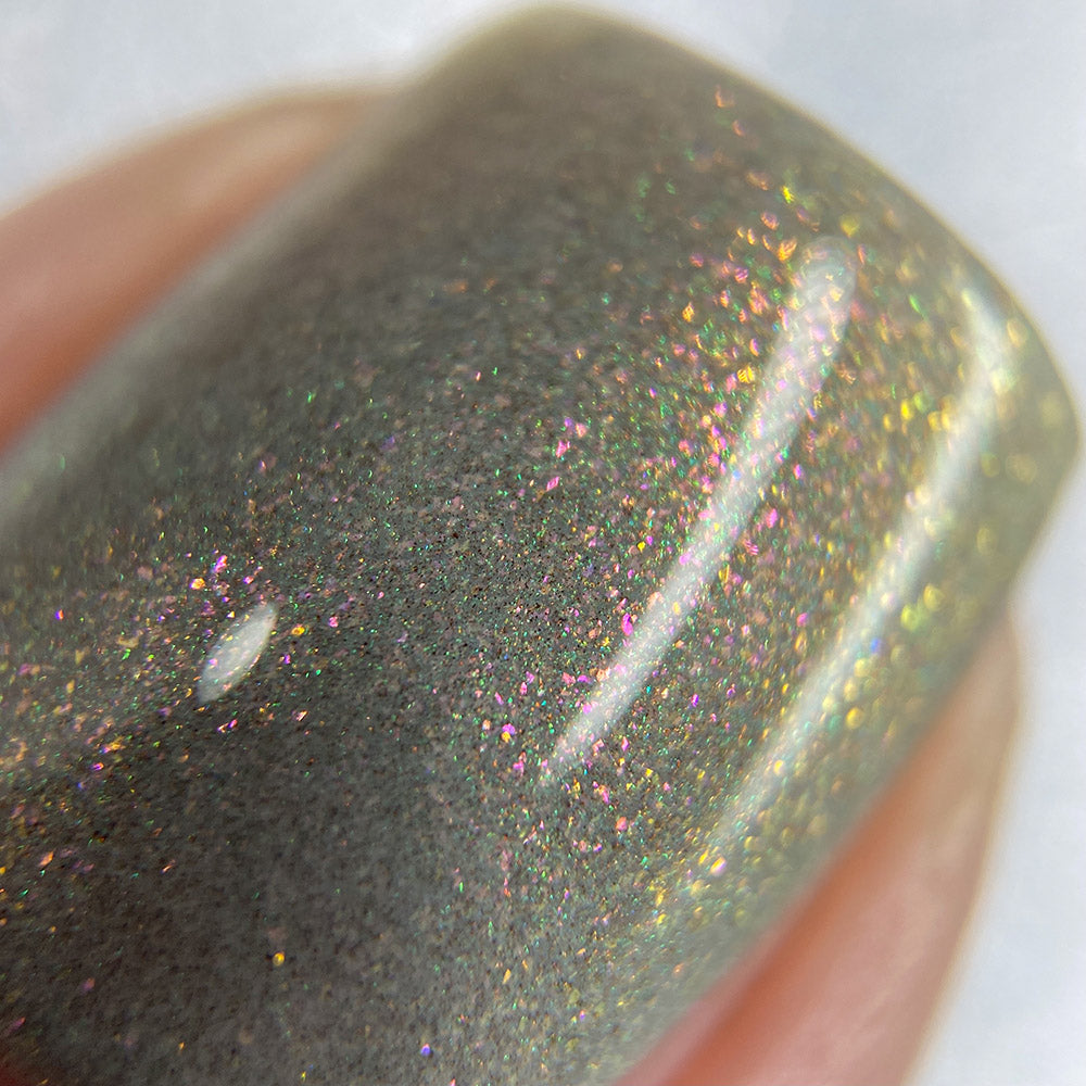 Close-up of a fingernail adorned with PI Colors' ToW.001 Taupe Nail Polish. This lacquer features a reflective, metallic finish with tiny multi-colored specks, creating an effect reminiscent of a shimmering galaxy that shifts subtly under the light. As a 10-free nail polish, it ensures a stunning look without harmful chemicals.