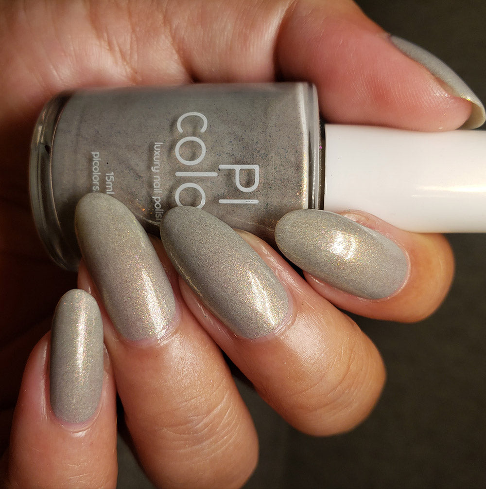 A hand with long nails painted in the shimmering shade of ToW.001 Taupe Nail Polish holds a matching bottle. The PI Colors bottle, featuring a white cap, showcases these 10-free nail polish nails meticulously manicured against a dark background.