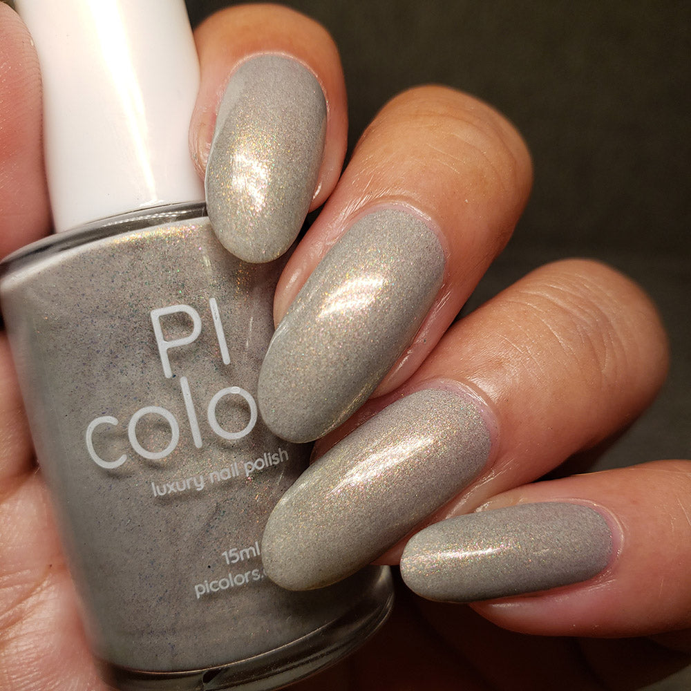 Close-up of a hand with long, oval-shaped nails painted in a shimmering taupe shade using "ToW.001 Taupe Nail Polish" by PI Colors. The person is holding a bottle of the same color polish labeled "PI Colors luxury 10-free nail polish.