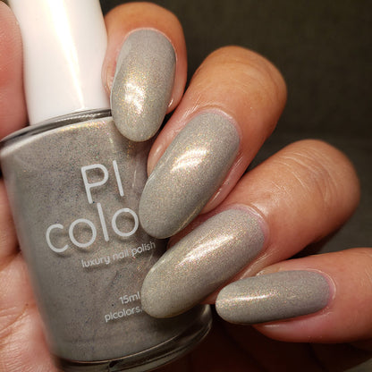 Close-up of a hand with long, oval-shaped nails painted in a shimmering taupe shade using "ToW.001 Taupe Nail Polish" by PI Colors. The person is holding a bottle of the same color polish labeled "PI Colors luxury 10-free nail polish.