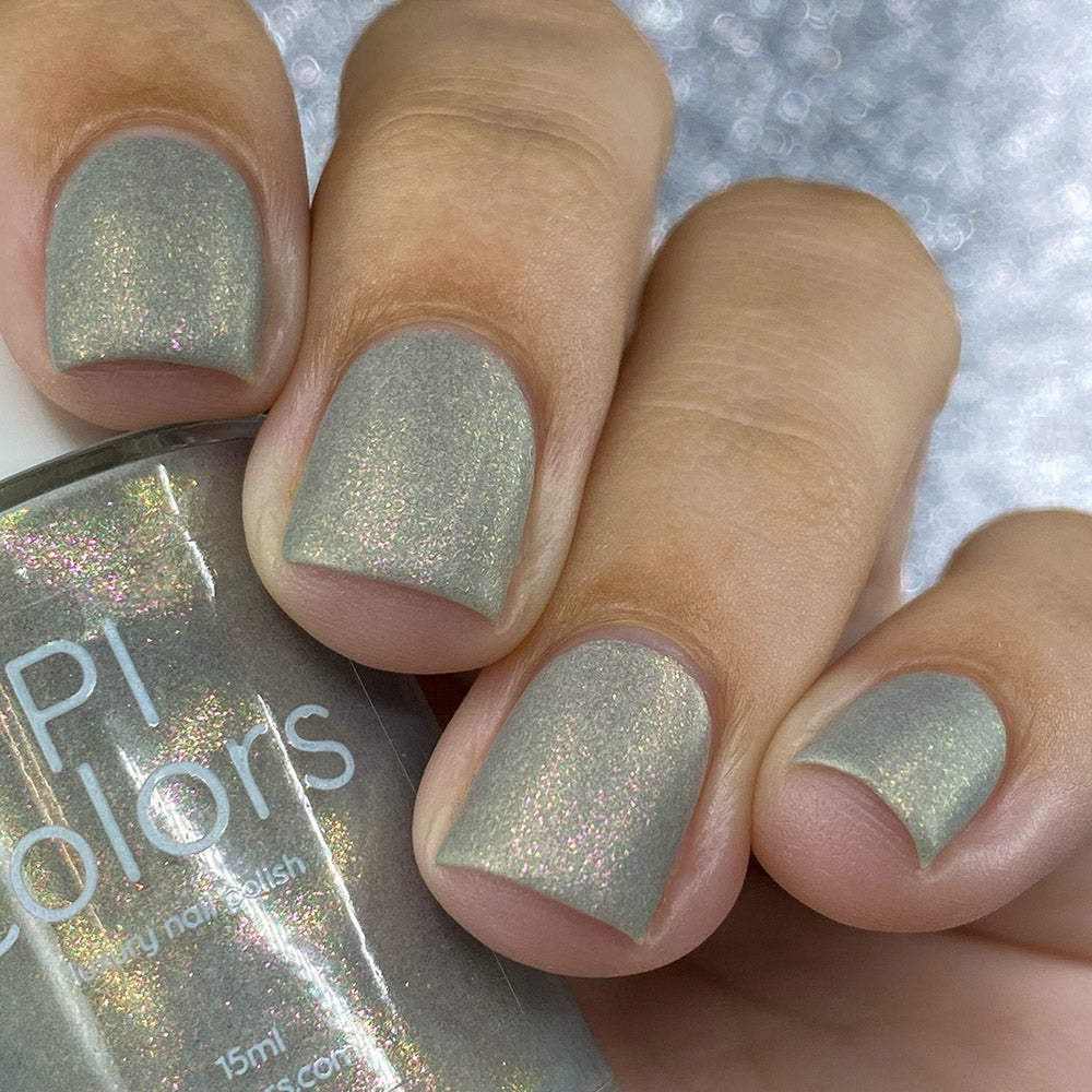 A close-up of a hand with short, well-groomed nails painted in PI Colors' ToW.001 Taupe Nail Polish, featuring a shimmering aurora finish with hints of pink glitter. The polish bottle is partially visible, held by the thumb in the foreground against a softly blurred background.