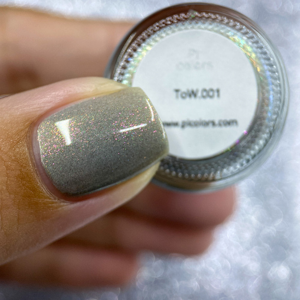 Close-up of a person holding a small jar of PI Colors ToW.001 Taupe Nail Polish against a sparkly background. Their thumb is adorned with shimmering aurora lacquer in a shimmery gray that perfectly matches the jar.