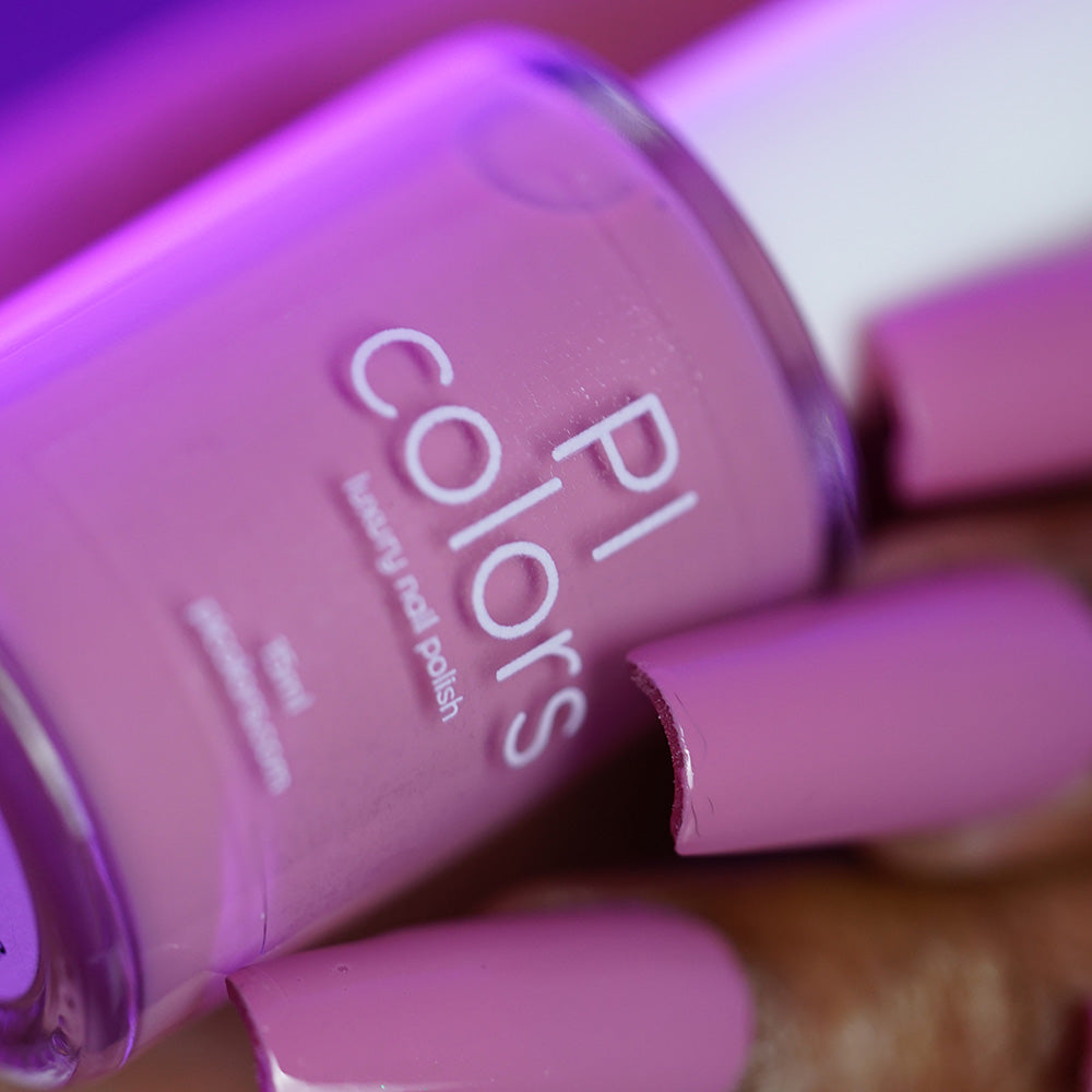 A close-up of a hand with long nails painted in PI Colors’ Pink Biscotti.462, a glossy pink-lavender creme polish, holding its matching bottle under purple lighting.