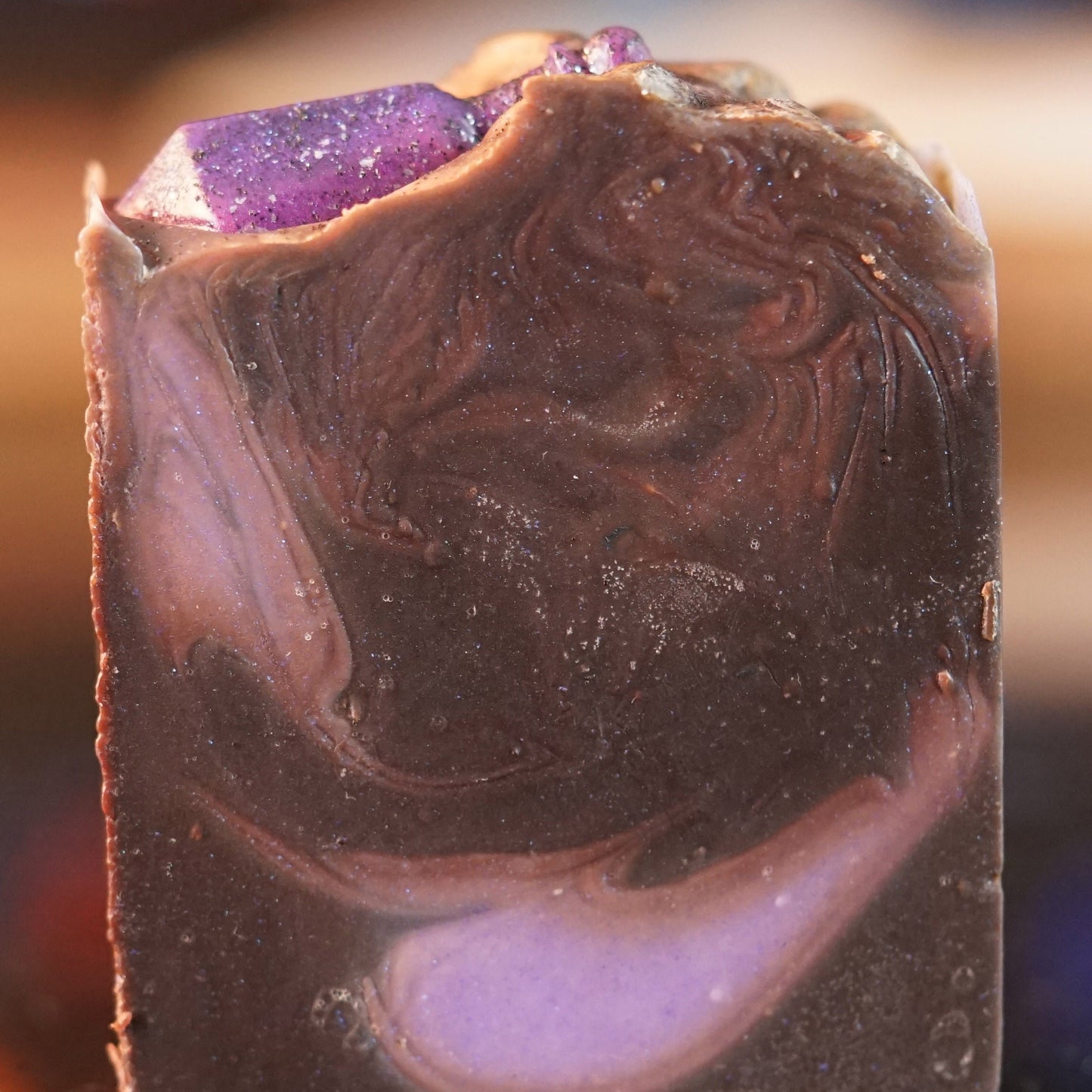 The Potion Drip Artisan Soap Bar by PI Colors showcases a glossy, marbled texture with swirls of dark brown and purple. A sparkly purple stone-like decoration crowns the rustic bar, offering an artisanal touch and promising a scented bath experience.