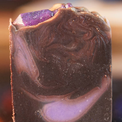 The Potion Drip Artisan Soap Bar by PI Colors showcases a glossy, marbled texture with swirls of dark brown and purple. A sparkly purple stone-like decoration crowns the rustic bar, offering an artisanal touch and promising a scented bath experience.