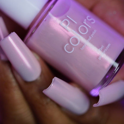 A close-up of a hand with long, light pink, glossy nails holds a bottle of PI Colors Snickerdoodle.405 handmade nail polish in a matching shimmery pale pink shade.