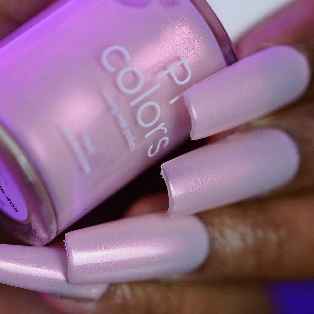 A hand with long, square-shaped nails is painted in PI Colors' Snickerdoodle.405, a shimmery pale pink nail lacquer with a delicate purple tint, holding the matching bottle under soft lighting.