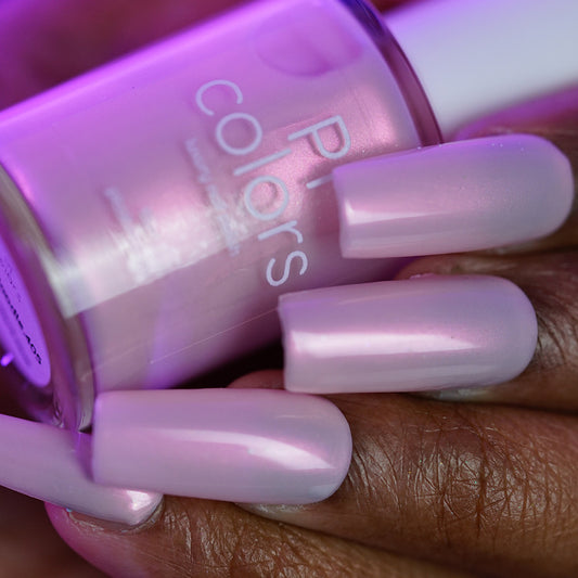 Close-up of a hand with long, almond-shaped nails painted in PI Colors Snickerdoodle.405, an iridescent pale pink handmade nail polish, holding the matching bottle.