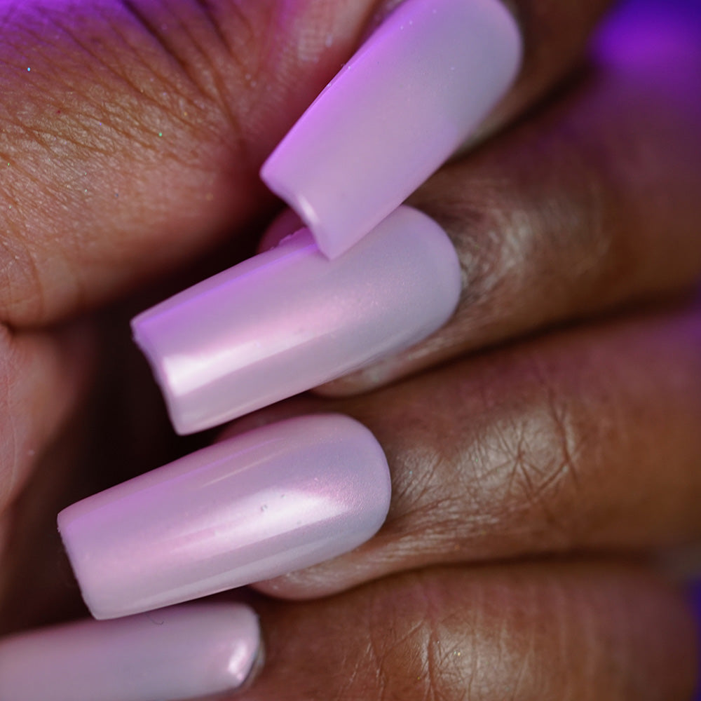 Close-up of a hand with long, square acrylic nails painted in PI Colors’ Snickerdoodle.405, a glossy pale pink polish with shimmer, against a purple-tinted background that highlights the luxurious finish on medium to dark skin.