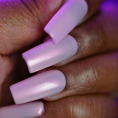 Close-up of a hand with long, square acrylic nails painted in PI Colors’ Snickerdoodle.405, a glossy pale pink polish with shimmer, against a purple-tinted background that highlights the luxurious finish on medium to dark skin.