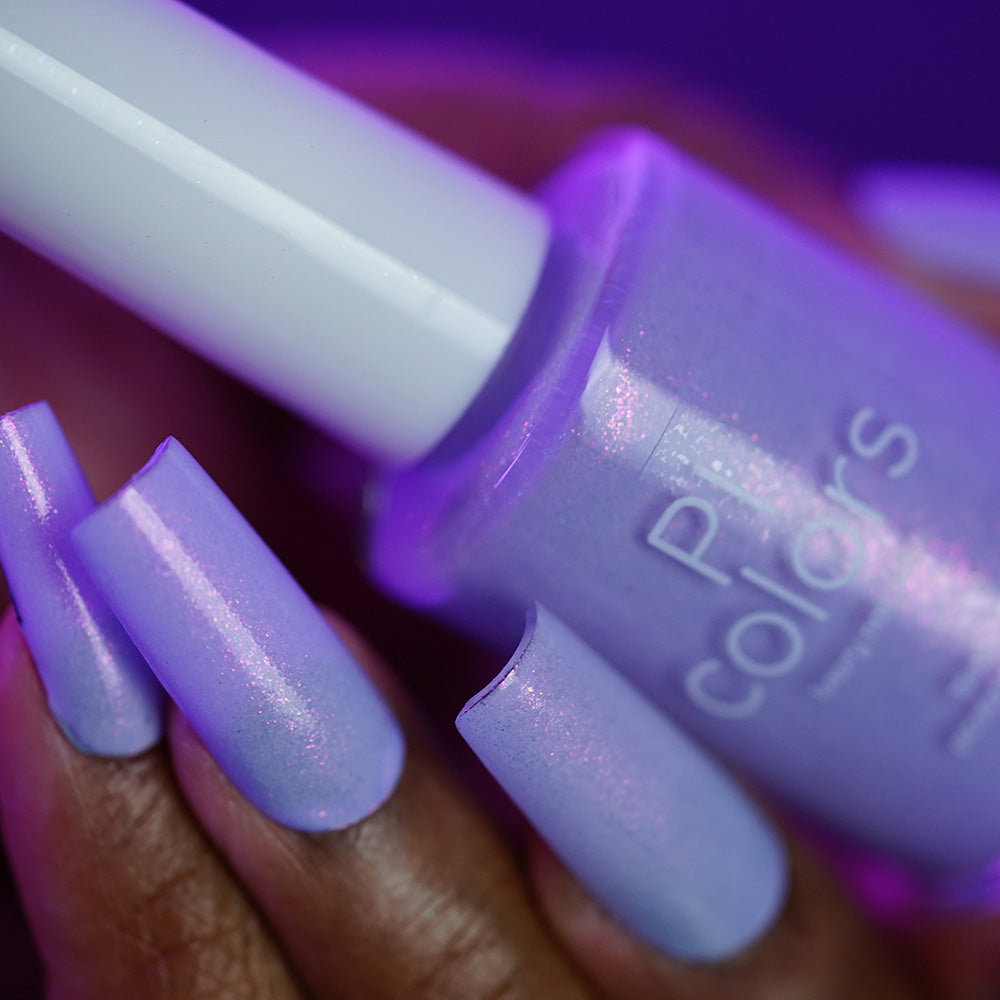 A hand with long, square nails painted in PI Colors Snowball.485—a shimmer lavender polish—holds the matching bottle with a white cap. The soft background features blurred purple tones.