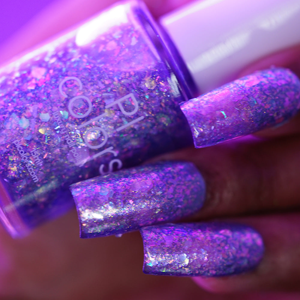 A close-up of a hand with long, purple glittery nails holding a matching bottle of PI Colors' Stained Glass.436 nail polish. Both shimmer with iridescent sparkles under bright light, highlighting this unique handmade polish.