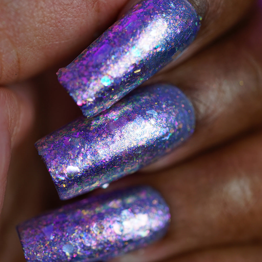 Fingernails on dark skin painted with PI Colors Stained Glass.436, an iridescent purple and blue flakie nail polish featuring shimmering flakes and a glossy finish.