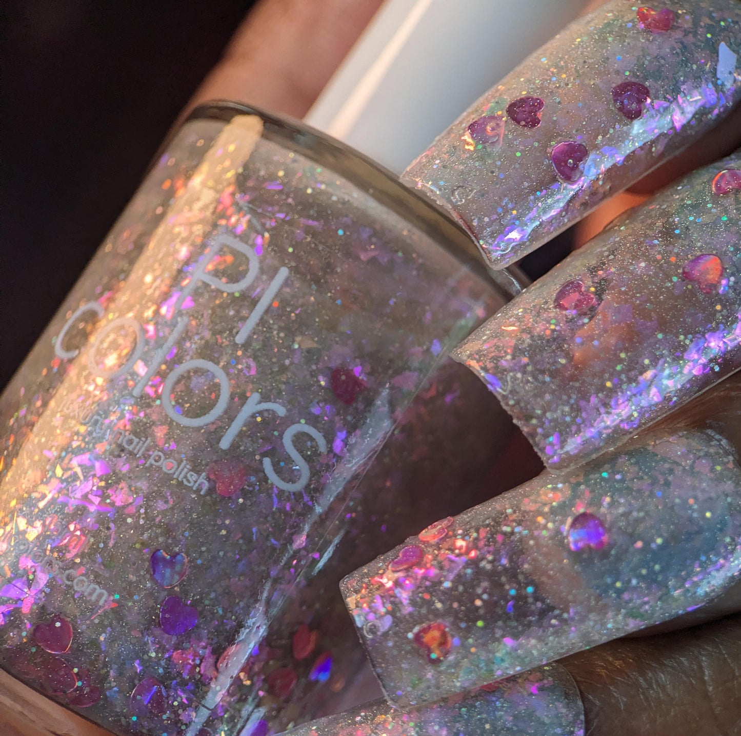 A close-up shows a hand with long nails painted in Stormy Hearts.206 by PI Colors, featuring a clear base with pink and holographic hearts, multicolored reflective flakes, and hints of silver holo glitter. The polished fingers hold a bottle of this dazzling topper.