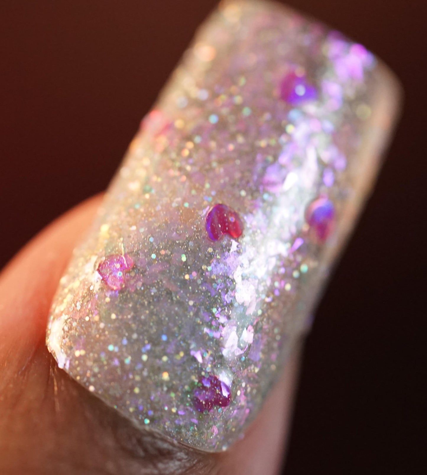 A close-up of a fingernail decorated with PI Colors Stormy Hearts.206 nail polish reveals glittery, translucent topper embellished with pink and purple circular confetti and shimmering holographic hearts, all set against a softly blurred background.