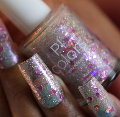 Close-up of a hand with long, square-shaped nails painted with Stormy Hearts.206 polish. It features silver holo glitter and multicolored holographic hearts. A partially visible nail polish bottle labeled "PI Colors" is in the background.