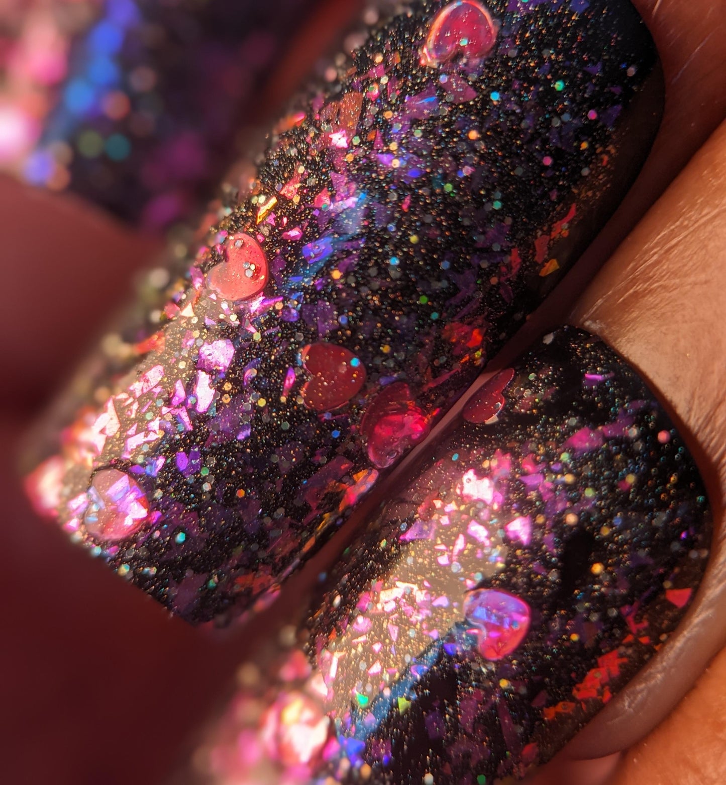 Close-up of three fingernails with PI Colors' Stormy Hearts.206 glossy black nail polish, topped with multicolored glitter and holographic hearts. The shimmering surface reflects a vibrant rainbow of colors.