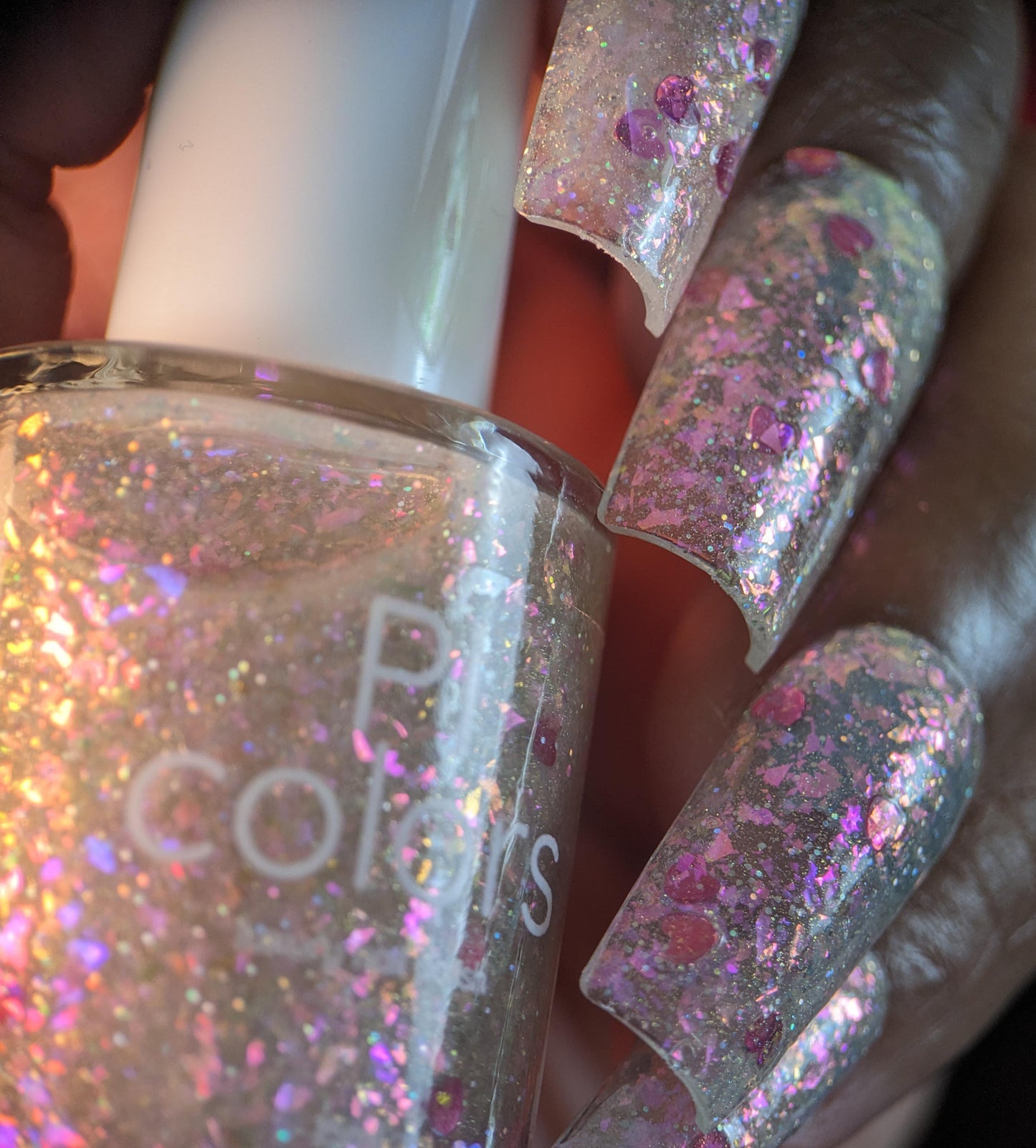 Close-up of a hand holding a PI Colors' "Stormy Hearts.206" nail polish bottle. The nails glisten with pink, purple, and silver sparkles, accented by holographic hearts. A white-capped bottle partially reveals its name amid the silver holo glitter twinkles.