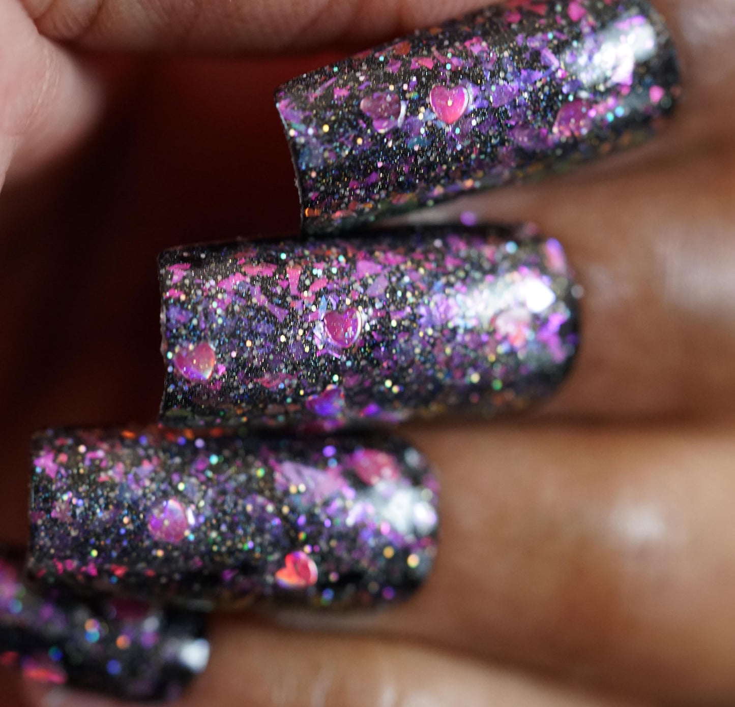 A close-up of fingers with long black nails showcases colorful glitter and silver holo glitter. Heart-shaped confetti enhances the charm, while Stormy Hearts.206 by PI Colors adds a shiny topper with pink and purple sparkles.