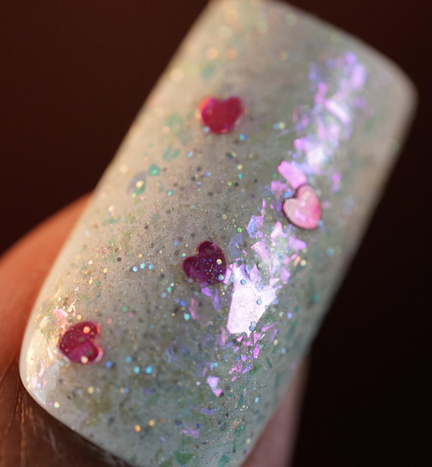 Close-up of a fingernail painted with PI Colors' Stormy Hearts.206 polish, showcasing a dazzling finish with multicolored glitter, holographic hearts, iridescent pink and purple flecks, and silver holo glitter for a shimmering effect.