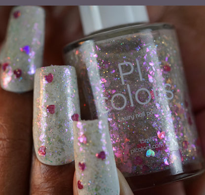 Close-up of fingers with long nails painted in a sparkly glitter nail polish featuring holographic hearts and pink accents. In the background, a bottle of PI Colors' Stormy Hearts.206 nail polish topper showcases a similar silver holo glitter design.