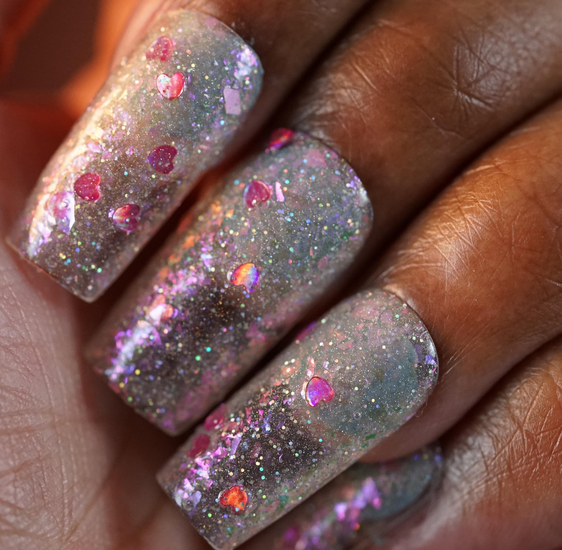 A close-up of a hand displays long nails with Stormy Hearts.206 nail polish from PI Colors, featuring a clear base and vibrant holographic hearts. Silver holo glitter adorns the nails, creating an eye-catching effect.