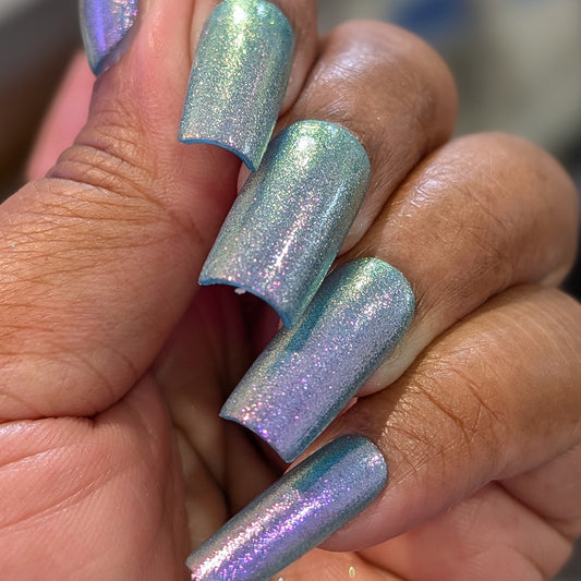 A close-up of a hand with long, square nails painted in PI Colors’ Sugar Sparkles.451, a luxury nail lacquer featuring shimmery, iridescent blue and purple hues with a metallic, holographic shine and an elegant finish.
